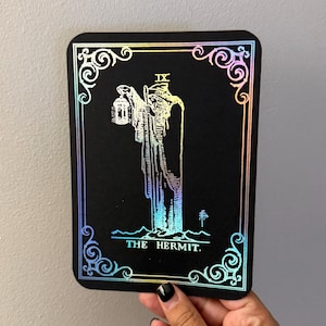 May include: A rectangular black card featuring a holographic foil illustration of The Hermit tarot card. The design includes the Roman numeral IX, a figure holding a lantern and staff, and ornate corner flourishes with the text THE HERMIT.
