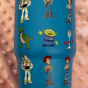 Puede incluir: Un vaso térmico azul con personajes de dibujos animados de Toy Story. El vaso tiene una tapa turquesa y está decorado con imágenes de Buzz Lightyear, Woody, Jessie, Rex, los Aliens y Forky.