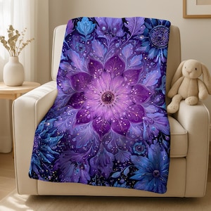Könnte beinhalten: Eine weiche Decke mit einem detaillierten Blumenmuster in Lila- und Blautönen. Die zentrale Blume ist ein leuchtendes Lila, mit aufwendigen Blütenblattdetails. Die Decke ist über einem cremefarbenen Sessel drapiert.