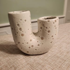May include: A speckled, off-white ceramic vase with a unique U-shaped design. The vase has two cylindrical openings connected by a curved body. The surface is textured with small, brown spots, adding visual interest to the decorative piece.