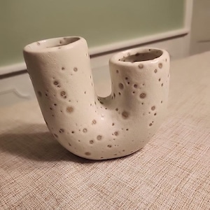 May include: A ceramic U-shaped vase with two openings. The vase is off-white with brown speckles. The vase is sitting on a textured, light brown surface.