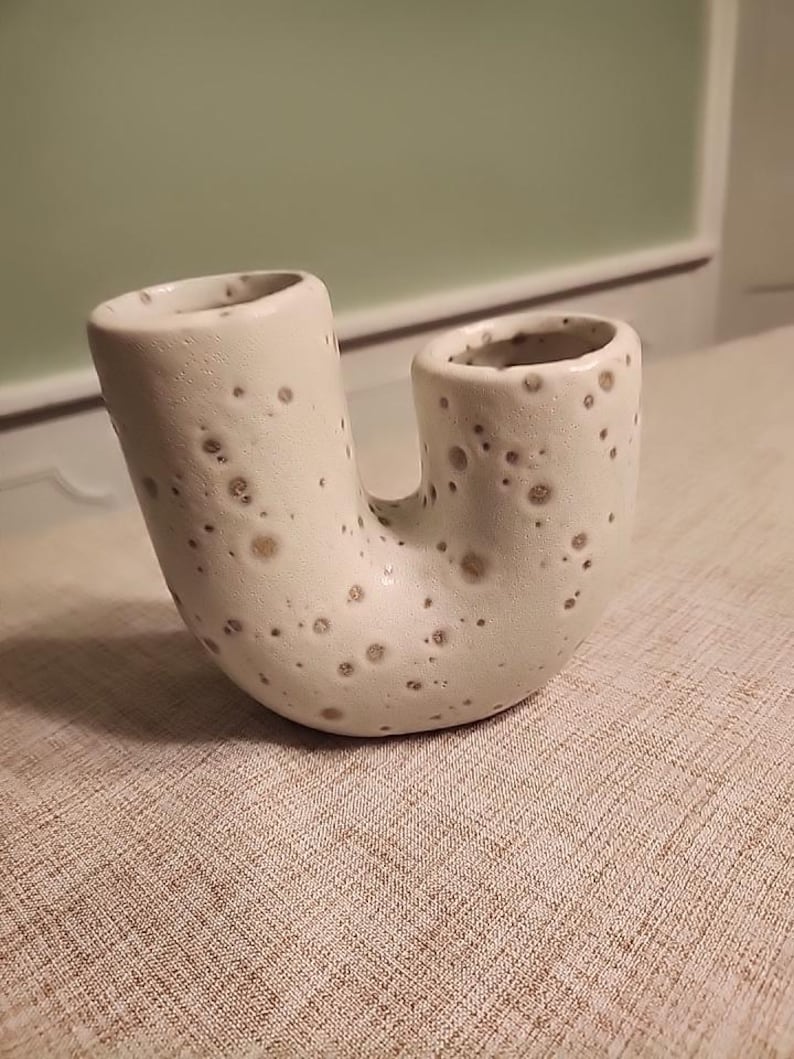 May include: A ceramic U-shaped vase with two openings. The vase is off-white with brown speckles. The vase is sitting on a textured, light brown surface.