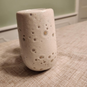 May include: A cylindrical ceramic cup with a speckled, off-white glaze. The cup has a rounded base and a smooth rim. The glaze features small, dark brown spots, giving it a unique, textured appearance. The cup is likely handmade and suitable for beverages.