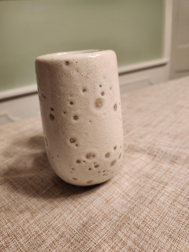 May include: A cylindrical ceramic cup with a speckled, off-white glaze. The cup has a rounded base and a smooth rim. The glaze features small, dark brown spots, giving it a unique, textured appearance. The cup is likely handmade and suitable for beverages.