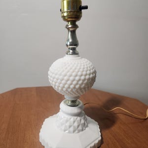 Puede incluir: Una lámpara de mesa blanca con diseño de clavos, con un casquillo de bombilla de latón y un vástago plateado. La lámpara tiene un globo redondo y texturizado y una base geométrica. La lámpara está sobre una mesa de madera.