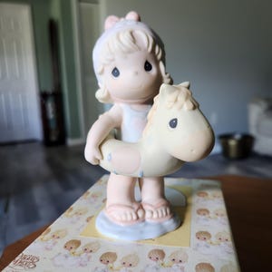 May include: A porcelain figurine of a child with blonde hair wearing a white hat and swimsuit, standing inside a cream-colored horse-shaped pool float with pink spots. The figurine stands on a box labeled Precious Moments Last Forever.