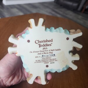 Puede incluir: Una figura de Cherished Teddies, con Jill, con el texto "I'm Always Good For A Soft Cuddle And A Warm Hug." La base es blanca con un borde festoneado azul claro y detalles de osito de peluche marr&oacute;n. Tambi&eacute;n est&aacute; impresa "First Edition".