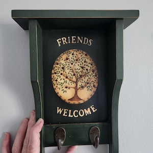 May include: A dark green wooden wall shelf with two hooks. The shelf features the words "FRIENDS WELCOME" above a painted tree design. The shelf is held by a hand.