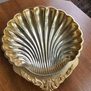 May include: A polished gold-toned metal dish shaped like a large scallop shell. The interior features deep, radiating fluted grooves, while the base is adorned with an ornate, raised decorative scrollwork pattern along the bottom edge.