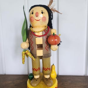 May include: A wooden nutcracker figurine featuring a painted face with black yarn hair and a feather. The figure wears a brown vest with red patterns, a gold and red beaded necklace, and holds green corn stalks and a round orange fruit.