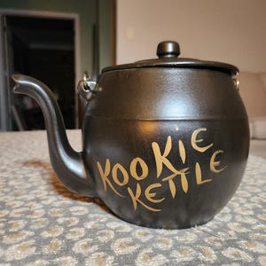 Puede incluir: Una tetera de cerámica negra con un pico curvo y una tapa. Las palabras "KOOKIE KETTLE" están escritas en letras doradas en la parte delantera de la tetera. La tetera está sobre una superficie estampada.