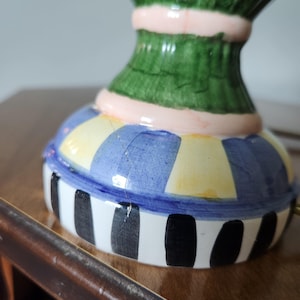 May include: A colorful ceramic decorative object with a base featuring blue, yellow, and white stripes. The object has a green upper section with a pink band. The base has black vertical stripes.
