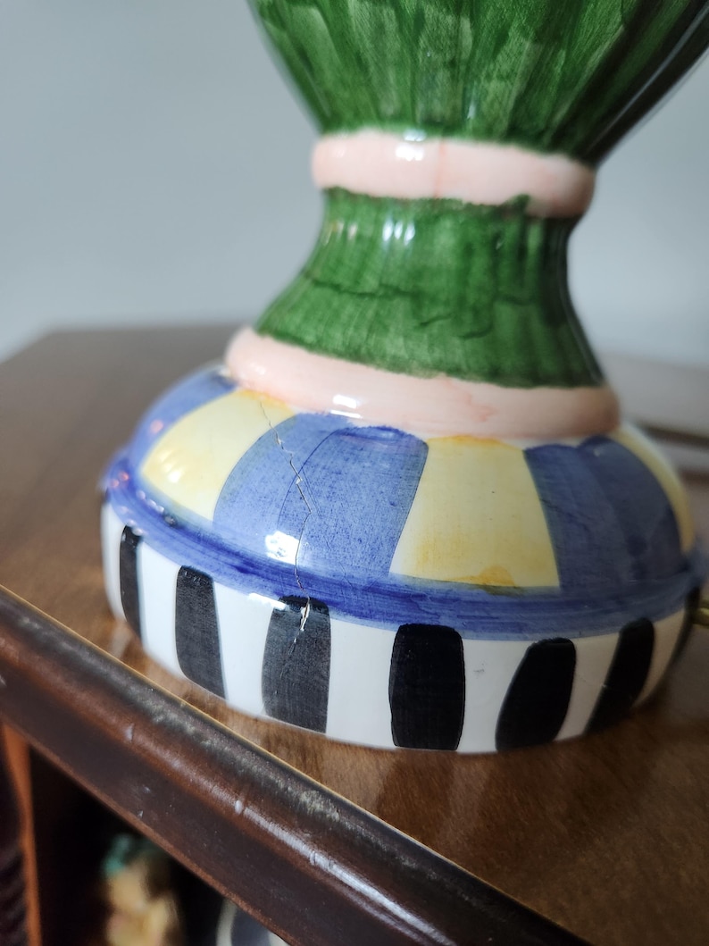 May include: A colorful ceramic decorative object with a base featuring blue, yellow, and white stripes. The object has a green upper section with a pink band. The base has black vertical stripes.