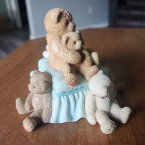 Puede incluir: Una figura de cuatro osos de peluche. Dos osos se abrazan sobre una almohada azul claro con un borde con volantes. Otros dos osos est&aacute;n sentados a cada lado. Los osos son de color canela y blanco con detalles en rosa.