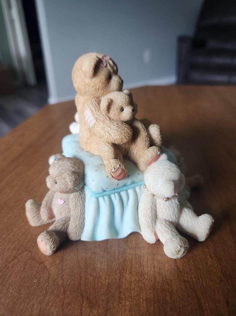 Puede incluir: Una figura de cuatro osos de peluche. Dos osos se abrazan sobre una almohada azul claro con un borde con volantes. Otros dos osos est&aacute;n sentados a cada lado. Los osos son de color canela y blanco con detalles en rosa.