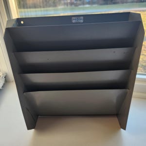 May include: A grey metal wall organiser with four angled compartments. The organiser has a rectangular shape and a small hole at the top. The brand name "HON" is visible on the top right corner. Designed to hold documents or other items.