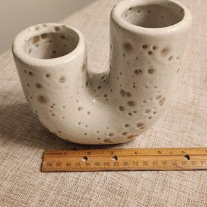 May include: A speckled, off-white ceramic vase with two cylindrical openings connected by a curved base. The vase has a unique, U-shaped design. A wooden ruler is placed next to the vase.