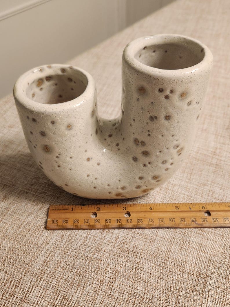 May include: A speckled, off-white ceramic vase with two cylindrical openings connected by a curved base. The vase has a unique, U-shaped design. A wooden ruler is placed next to the vase.