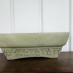 May include: A rectangular ceramic planter in a pale sage green color with a speckled finish. The base of the planter features a raised geometric pattern consisting of repeating angular, stepped motifs that wrap around the lower section.