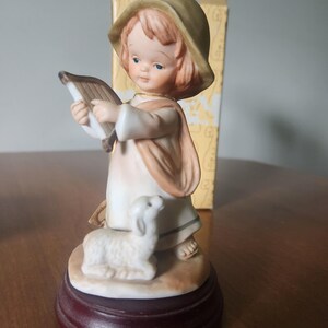 May include: A porcelain figurine of a child wearing a tan hat and a light-coloured robe, playing a small harp. A white lamb sits at the child's feet on a tan base. The entire figurine is mounted on a circular dark brown wooden pedestal base.