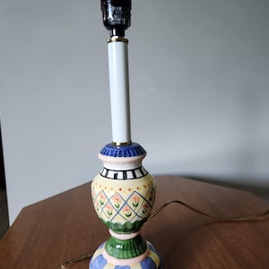 May include: A decorative table lamp with a white stem and a colorful base. The base features a yellow, green, blue, and black design with floral and geometric patterns. The lamp has a black lamp holder and a gold accent.