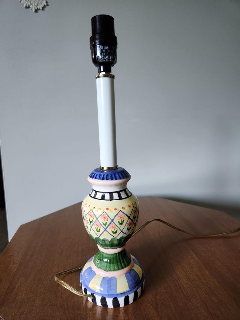 May include: A decorative table lamp with a white stem and a colorful base. The base features a yellow, green, blue, and black design with floral and geometric patterns. The lamp has a black lamp holder and a gold accent.