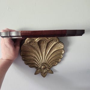 May include: A wall-mounted shelf featuring a dark brown wooden top surface and a decorative gold-toned metal bracket shaped like a large, detailed scallop shell. The bracket has a fan-like design with vertical ridges and a small floral base.