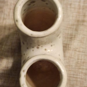 May include: A cream-colored ceramic pipe-shaped vase with two circular openings. The vase has a speckled pattern and a curved design. The interior of the vase is a warm brown color.