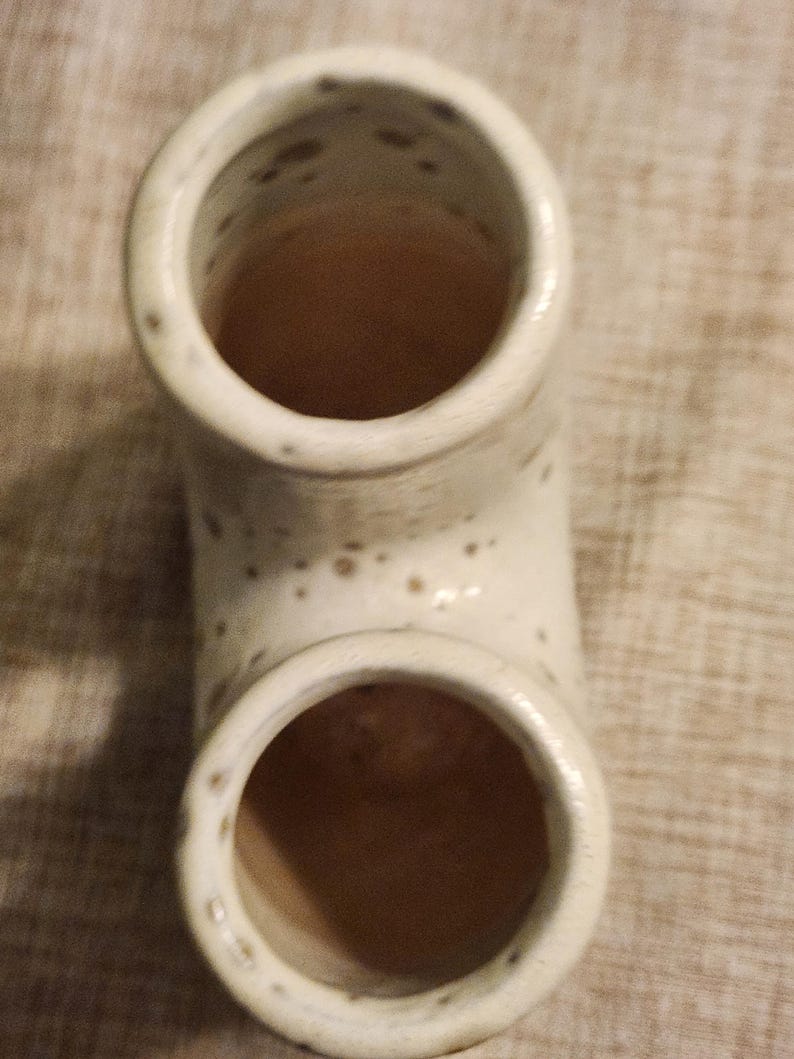 May include: A cream-colored ceramic pipe-shaped vase with two circular openings. The vase has a speckled pattern and a curved design. The interior of the vase is a warm brown color.