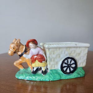May include: A ceramic figurine depicting a horse pulling a cart with a person. The horse is brown and the cart is white with a basket weave design. The person is wearing a red hat and pink shirt. The base is green.