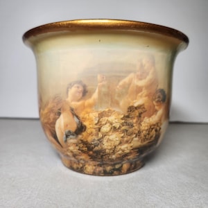 May include: A ceramic planter with a cream-colored base and a metallic gold rim. The exterior features a detailed transfer print depicting several cherubs or putti figures surrounded by clusters of grapes and foliage in soft, muted earth tones.