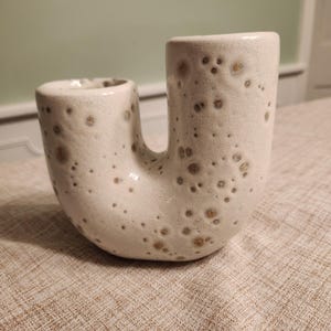 May include: A cream-colored ceramic vase with a unique U-shape design. The vase has a textured surface with brown speckles. The vase is a decorative item, suitable for displaying flowers or as a standalone art piece.