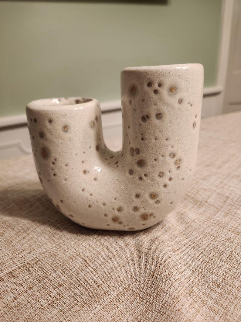 May include: A cream-colored ceramic vase with a unique U-shape design. The vase has a textured surface with brown speckles. The vase is a decorative item, suitable for displaying flowers or as a standalone art piece.