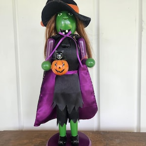 May include: A green-skinned witch nutcracker figure wearing a black hat with an orange band, a black dress with a jagged hem, and a purple-lined cape. The figure has long brown hair, holds a small orange pumpkin with a black cat, and stands on a purple base.