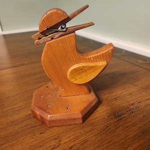 Puede incluir: Un soporte de pinza de ropa con forma de pájaro de madera. El pájaro está hecho de madera marrón claro con un pico articulado y un ala pequeña de color más claro. La base tiene forma octogonal. La pinza de ropa se sujeta con un muelle metálico.
