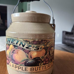Könnte beinhalten: Ein Vintage-Steinguttopf von Heinz's Apple Butter mit schwarzem Griff und Metalldraht. Das Etikett zeigt Illustrationen von Früchten wie Äpfeln, Birnen und Pflaumen. Der Topf ist beige mit einem Deckel.