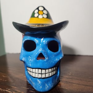 May include: A blue ceramic skull figurine featuring a distressed paint finish and a wide-brimmed hat. The hat is decorated with a yellow band and a white flower pattern with a yellow center. The skull has hollow eye sockets and a painted white toothy grin.