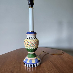 May include: A colorful ceramic table lamp with a black lamp holder and a white column. The base features a yellow, green, blue, and black design with floral and geometric patterns. The lamp has a gold-colored cord.