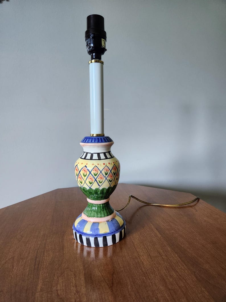 May include: A colorful ceramic table lamp with a black lamp holder and a white column. The base features a yellow, green, blue, and black design with floral and geometric patterns. The lamp has a gold-colored cord.