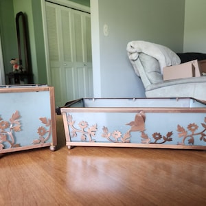May include: Two rectangular containers with frosted glass panels and copper-toned metal frames. Each piece features decorative copper-toned metal floral and leaf cutouts applied to the glass. Both containers stand on small, rounded metal feet.