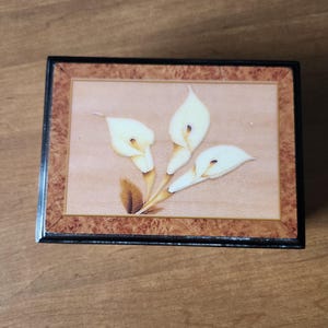 May include: A rectangular wooden music box with a floral design. The lid features a light brown background with three white calla lilies with yellow stems and a brown border. The box has a black frame.
