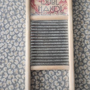 Op de afbeelding: Een vintage wasbord met een houten frame en een gegolfd metalen schrobvlak. Het bovenste gedeelte toont de tekst "DUBL HAND" in rood en zwart, met extra tekst eronder. Het wasbord is lichtbruin van kleur.
