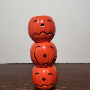 May include: A vertical stack of three orange ceramic pumpkin-shaped containers. Each pumpkin features a distinct black painted facial expression, including arched eyebrows, a small circular nose, and a wavy or curved mouth line, with a smooth finish.
