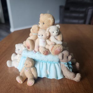 Puede incluir: Una figura decorativa con una colecci&oacute;n de ositos de peluche de varios tama&ntilde;os y poses. Los osos son principalmente de color marr&oacute;n claro y blanco, dispuestos sobre una base azul claro con un estampado de lunares y un borde con volantes. Algunos osos tienen corazones rosas.