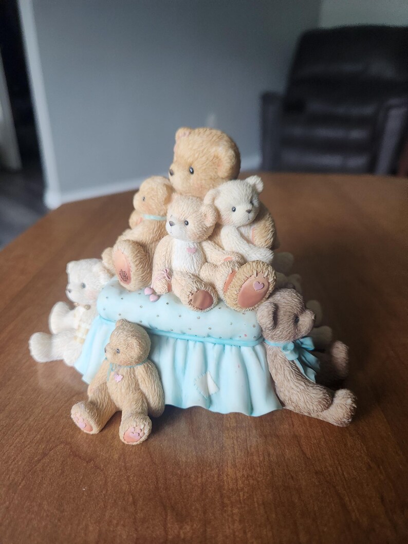 Puede incluir: Una figura decorativa con una colecci&oacute;n de ositos de peluche de varios tama&ntilde;os y poses. Los osos son principalmente de color marr&oacute;n claro y blanco, dispuestos sobre una base azul claro con un estampado de lunares y un borde con volantes. Algunos osos tienen corazones rosas.