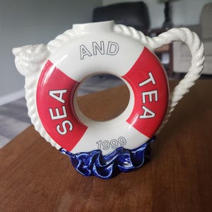 May include: A ceramic teapot shaped like a life preserver, with the words "SEA AND TEA 1999" in black lettering. The life preserver is white with a red stripe. The base is blue, resembling waves, and the handle is rope-like.