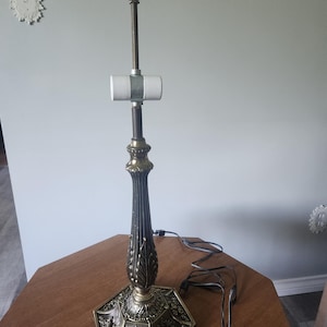 May include: An antique brass-toned lamp base with a hexagonal base and ornate floral detailing. The lamp's central column has a fluted design and a bulb socket. A power cord extends from the base.
