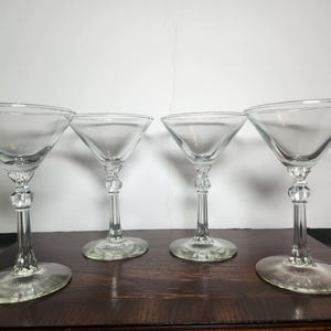 May include: A set of four clear glass martini glasses, each featuring a wide, conical bowl, a slender stem with a decorative spherical node, and a circular base. The glassware is transparent and smooth, designed with a classic silhouette.