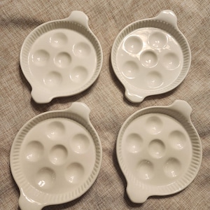 May include: Four white ceramic escargot plates. Each plate has a circular shape with indentations for the snails and a small handle. The plates have a ridged border.