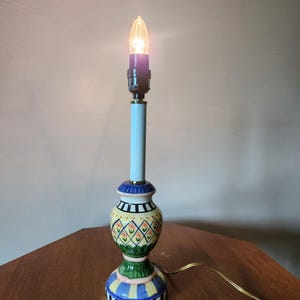 May include: A decorative table lamp with a lit bulb. The lamp has a light blue stem and a colorful base with yellow, green, blue, and black patterns. The lamp is on a wooden surface.